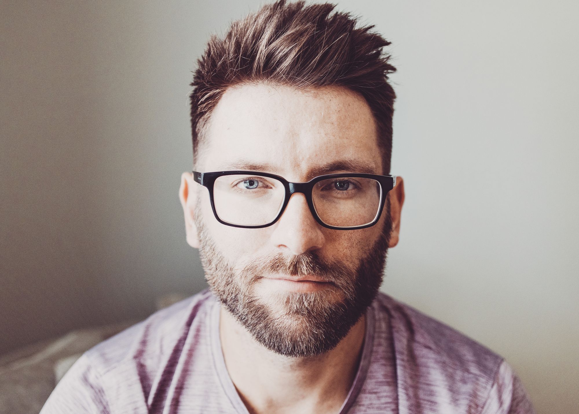 Headshot of mid-30s man with short brown hair, groomed beard, black glasses and a purple workout shirt sitting in a chair and smirking